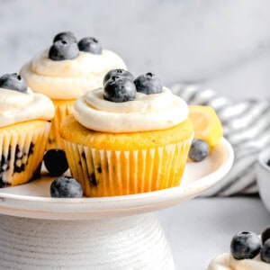 lemon blueberry cupcake on a cake platter next to two other cupcakes and fresh blueberries and lemons
