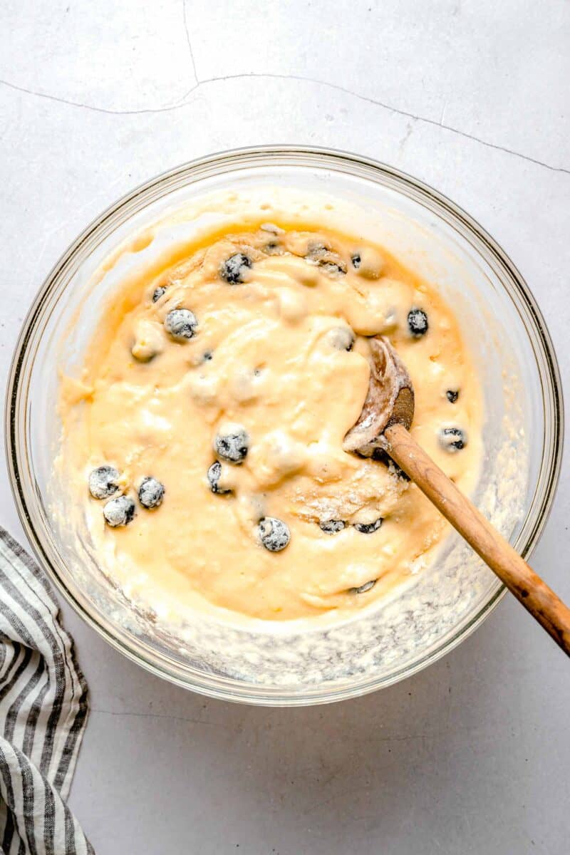 blueberries in the lemon blueberry cupcake batter in a clear bowl with a wooden spoon