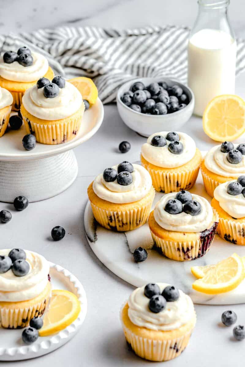 lemon blueberry cupcakes on marble plates and cake platters with fresh blueberries in a bowl and a milk jug