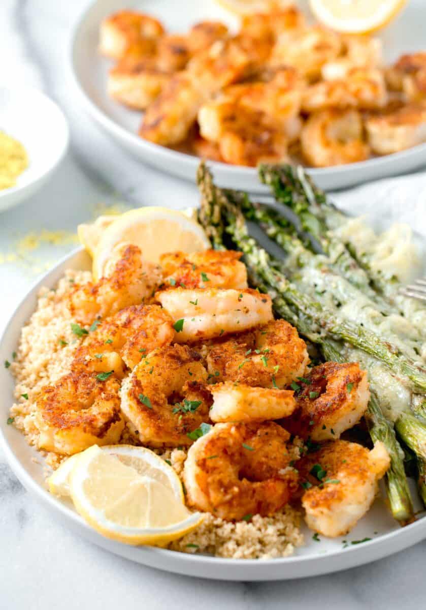 lemon pepper shrimp on top of couscous and next to parmesan crusted asparagus on a white plate with fresh lemon slices