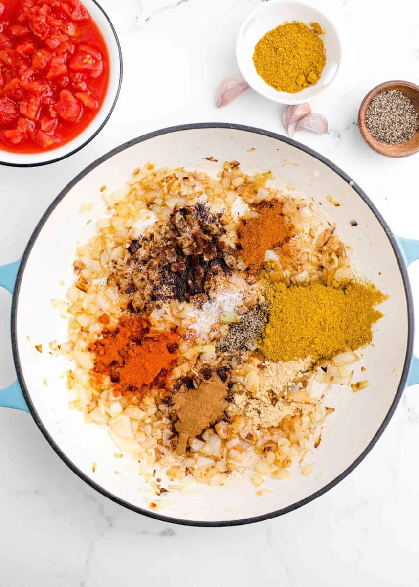 diced onions and spices in a cast iron skillet next to a bowl of tomatoes, garlic, spices, and black pepper