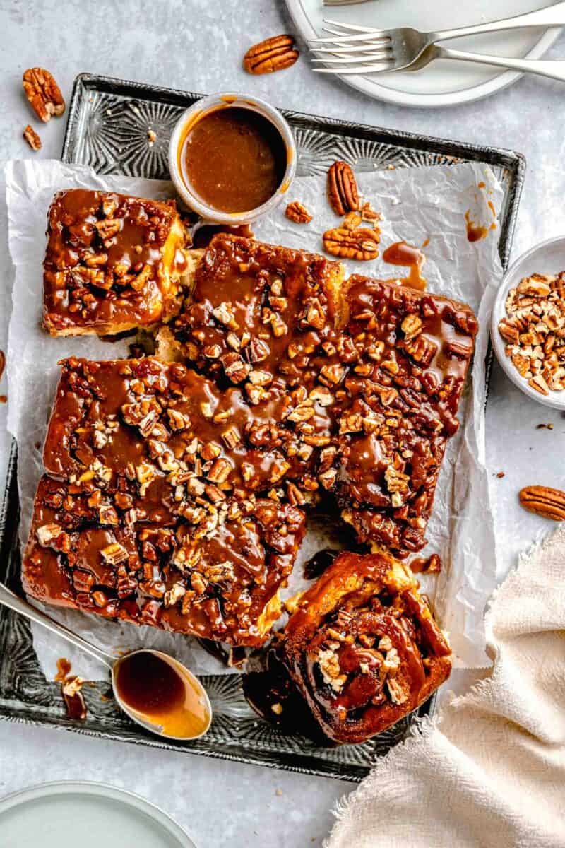 sticky buns and glaze on top of a metal baking sheet with a linen towel and a bowl of chopped pecans next to the baking sheet