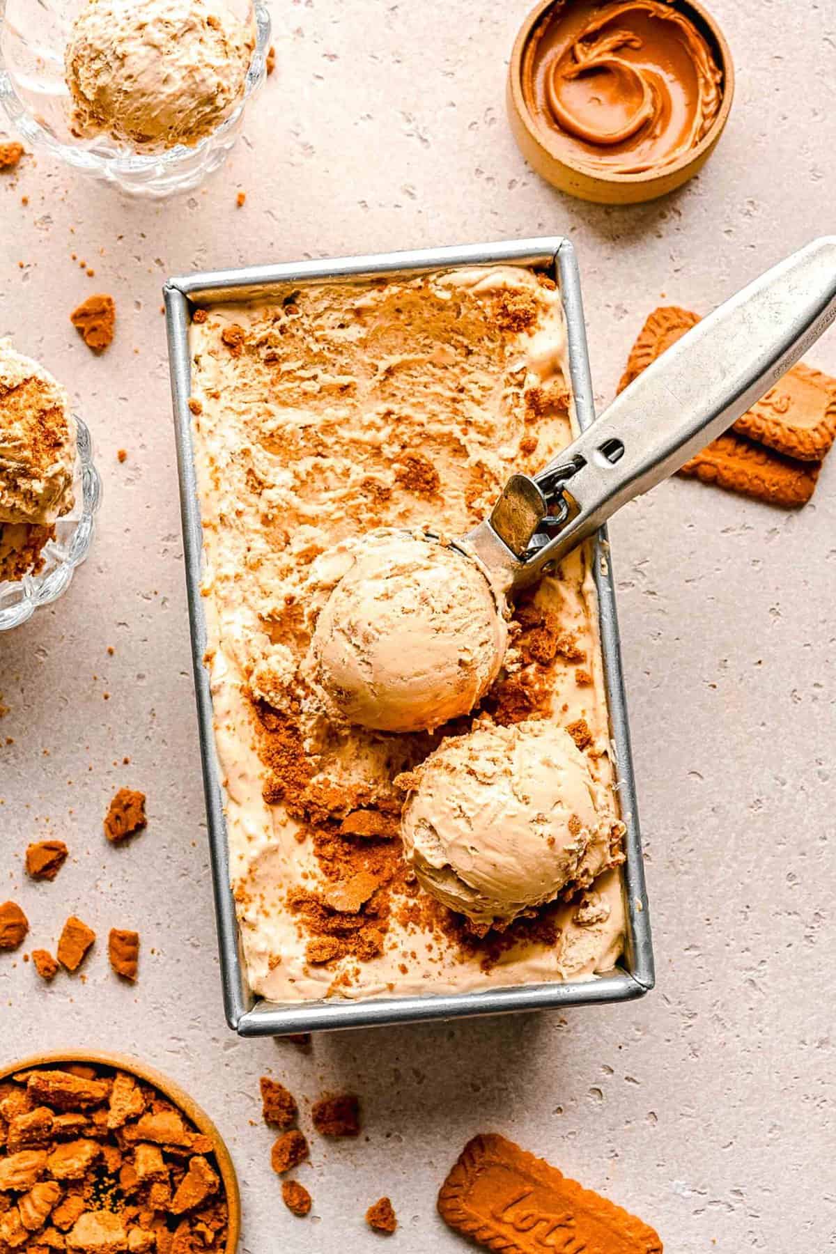 Overhead image of an ice cream scoop scooping Biscoff ice cream out of a loaf pan surrounded by Biscoff cookies and cookie spread.