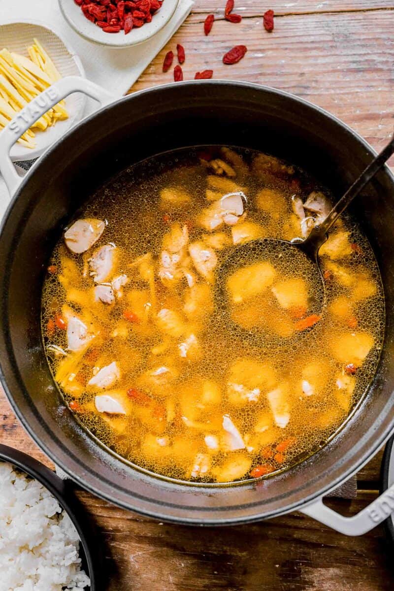 chinese chicken noodle soup in a large pot with ladle in the soup. small bowls of ginger, goji berries, and white rice can be seen around the pot.