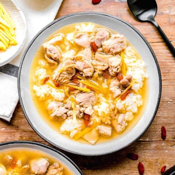 drunken chinese chicken soup with white rice in a white ceramic bowl next to smaller bowls of ingredients and a metal black spoon