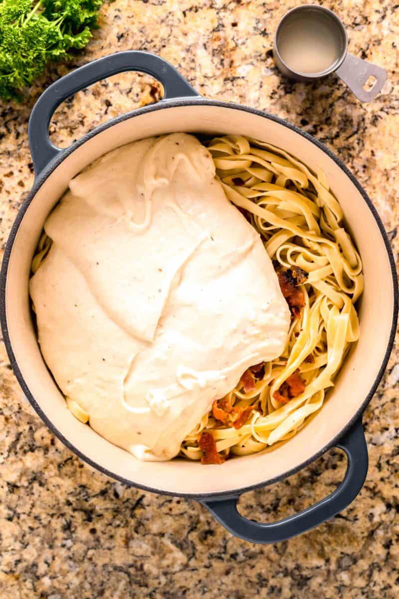 cream sauce poured on top of pasta and bacon in a blue dutch oven