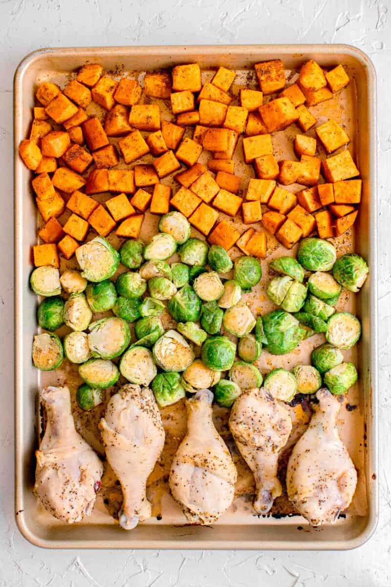 uncooked sweet potatoes, brussels sprouts, and chicken legs lined evenly on a rimmed baking sheet