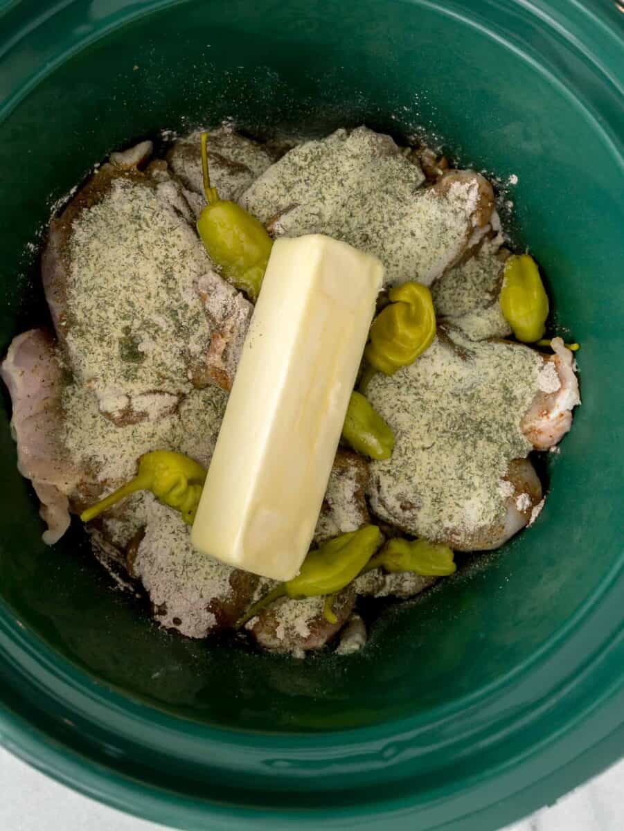 chicken in a green ceramic slow cooker insert with au jus gravy mix and ranch seasoning sprinkled on top along with pepperoncinis and a stick of butter