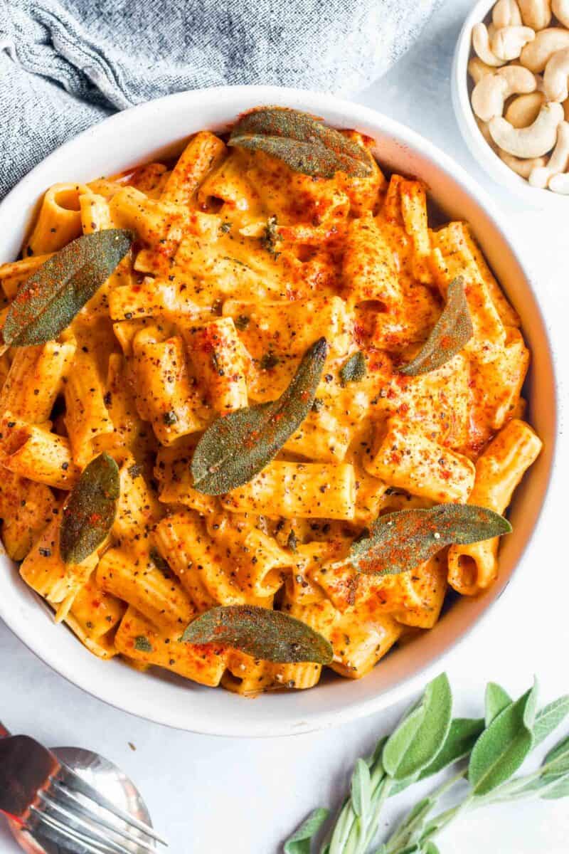 rigatoni pasta in a creamy pumpkin sauce topped with sage and fresh paprika and ground black pepper in a white bowl next to fresh sage and a metal fork