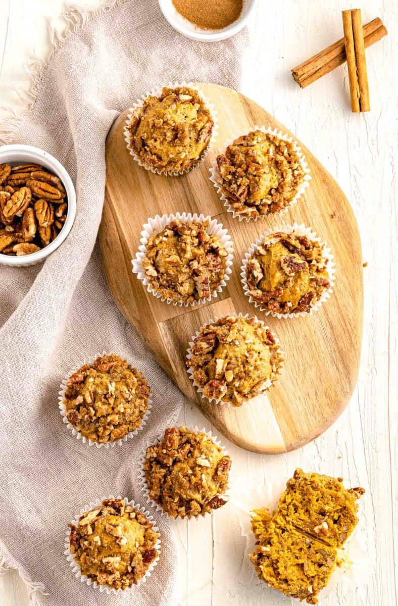 pumpkin pecan muffins on top of a wooden cutting board next to a brown linen towel and cinnamon sticks and spices