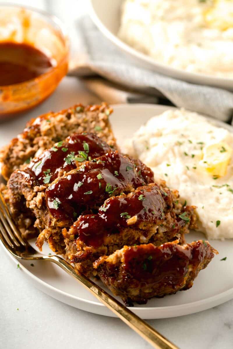 meatloaf slices on a large white rimmed plate with mashed potatoes on the side and a gold metal fork