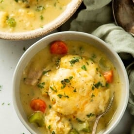 a grey bowl next to another bowl both filled with chicken and dumplings soup and a big cheddar biscuit on top
