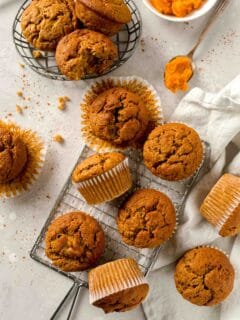 Overhead view of pumpkin banana muffins on a wire cooling rack next to more muffins in a basket and a spoonful of pumpkin puree.