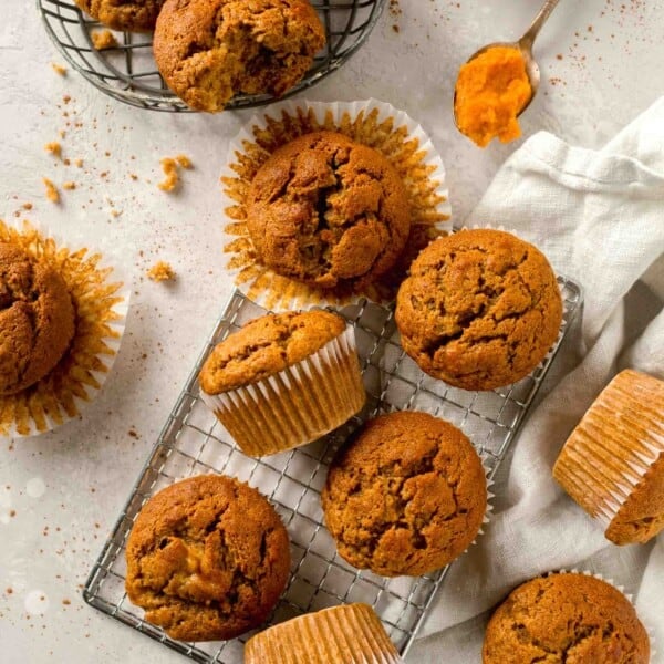 Overhead view of pumpkin banana muffins on a wire cooling rack next to more muffins in a basket and a spoonful of pumpkin puree.