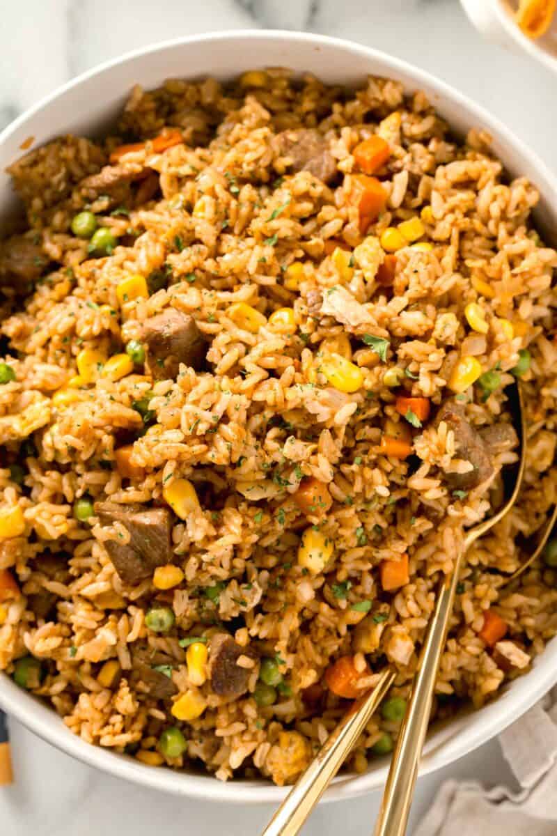 an overhead image of steak fried rice in a large white bowl with two spoons in the bowl