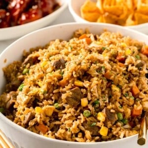 a large bowl of steak fried rice next to fried wontons and wooden chopsticks