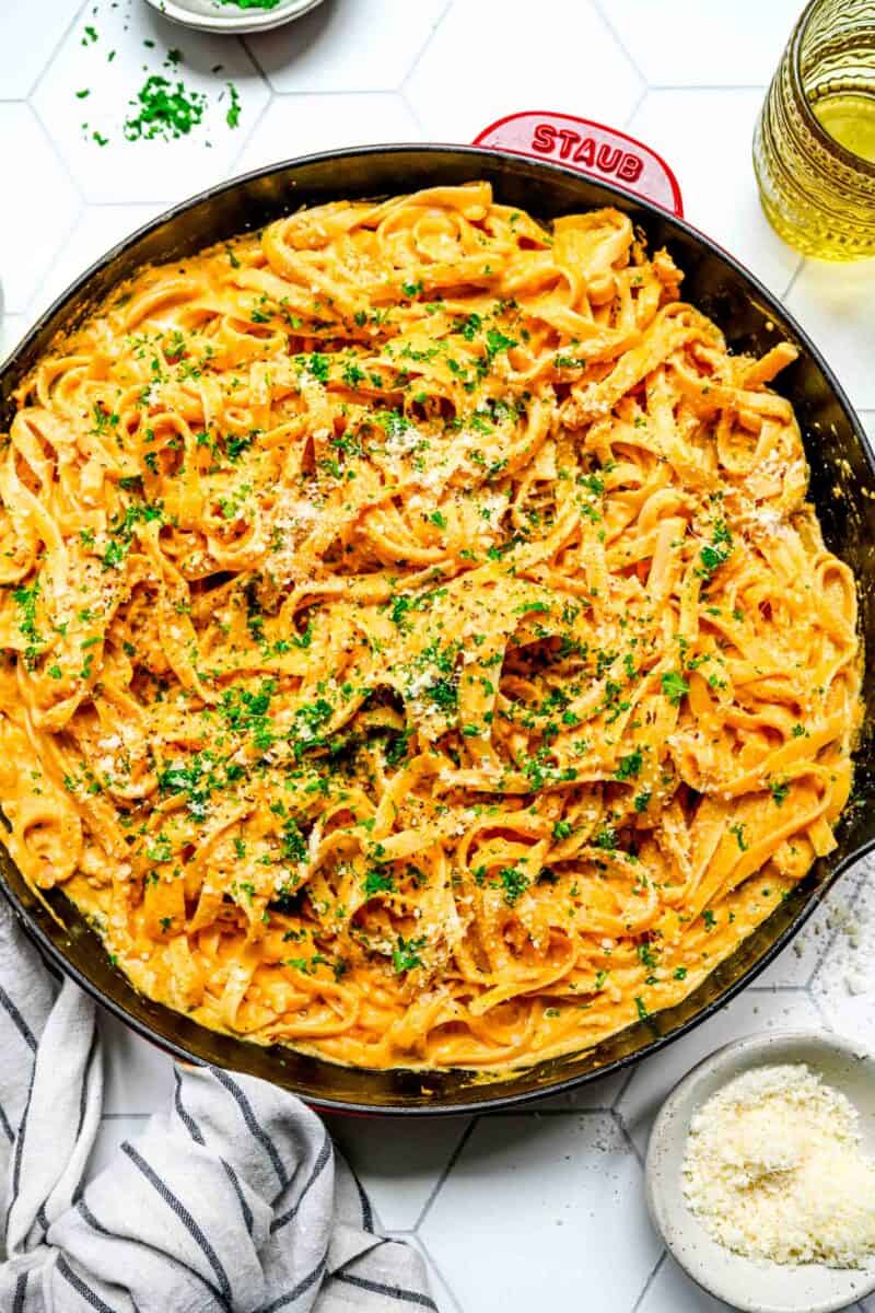 pumpkin alfredo pasta topped with fresh parsley in a red cast iron skillet next to a green glass and linen towel