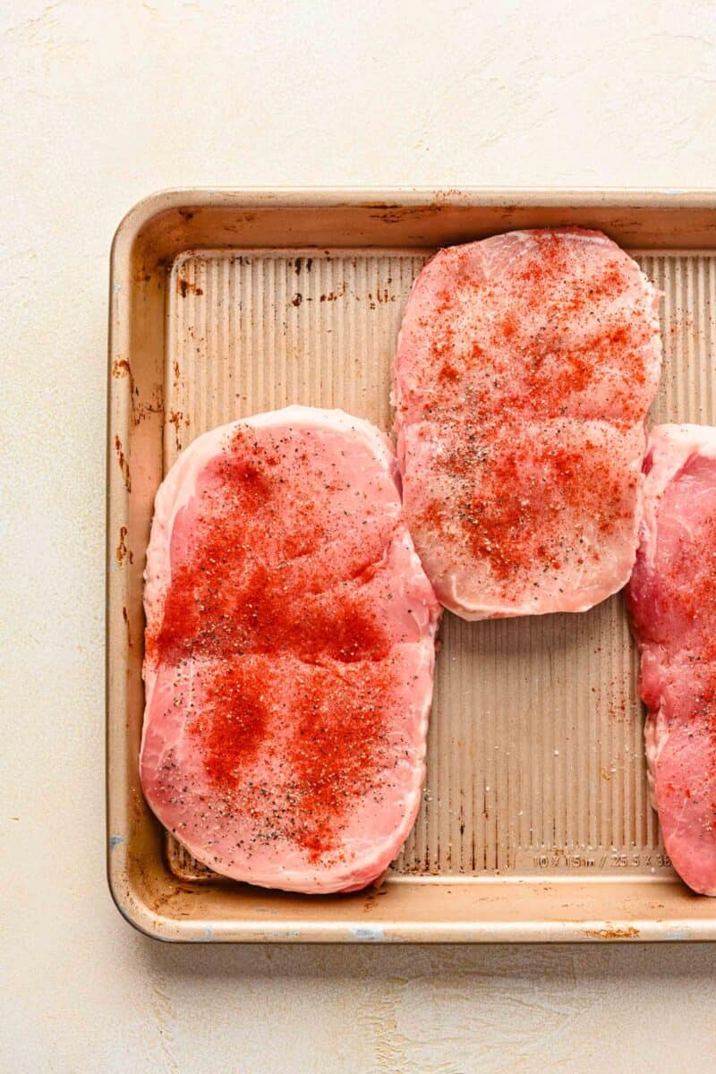 raw pork chops on a metal sheet pan with seasonings sprinkled on top