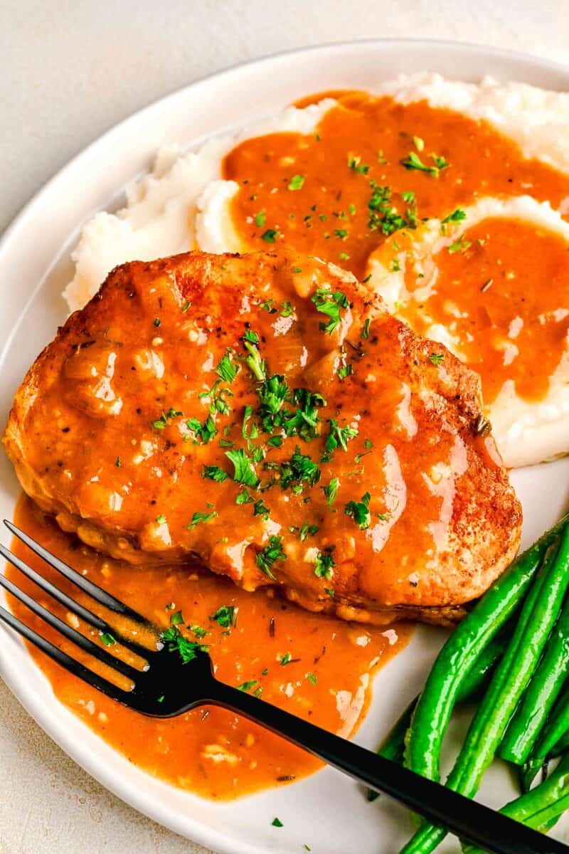 pork chops next to mashed potatoes and green beans with pan drippings made into a gravy on top