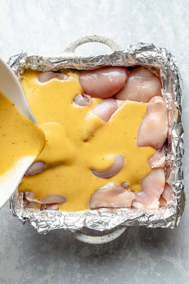 Pouring sauce over raw chicken thighs in a baking dish lined with aluminum foil.