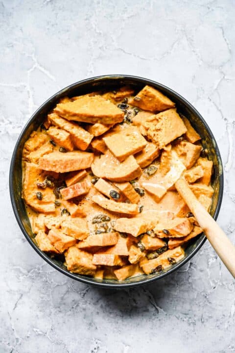a spatula stirs together bread pieces, custard mix, and raisins