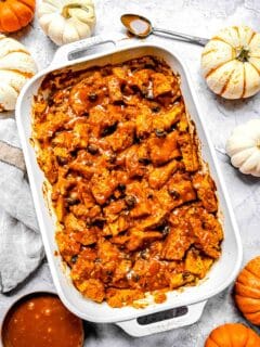 baked pumpkin bread pudding a white casserole dish with salted caramel drizzle on top. there are pumpkins arranged around the casserole dish