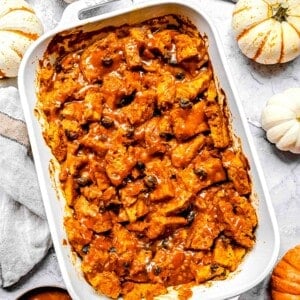 baked pumpkin bread pudding a white casserole dish with salted caramel drizzle on top. there are pumpkins arranged around the casserole dish