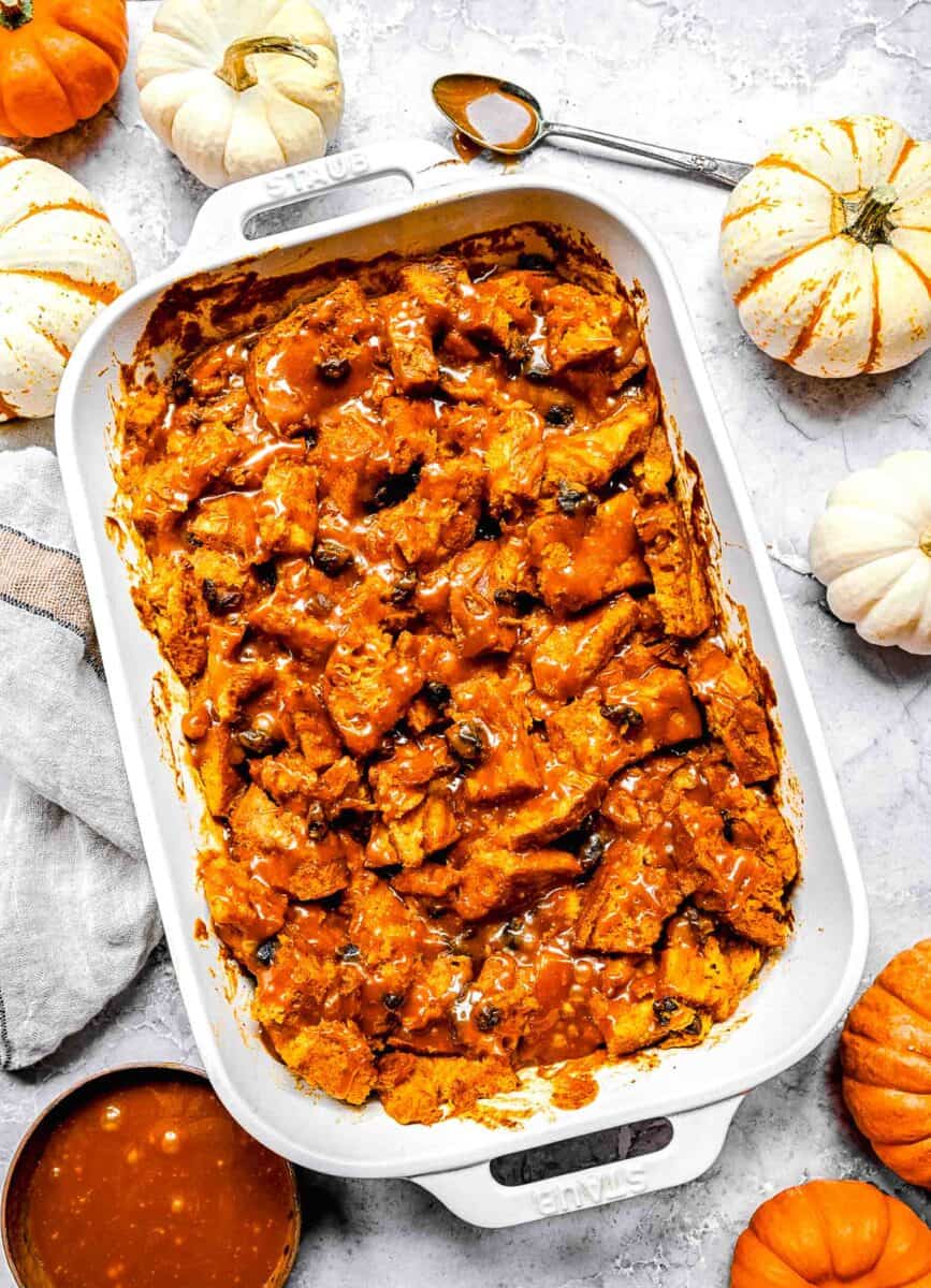 baked pumpkin bread pudding a white casserole dish with salted caramel drizzle on top. there are pumpkins arranged around the casserole dish