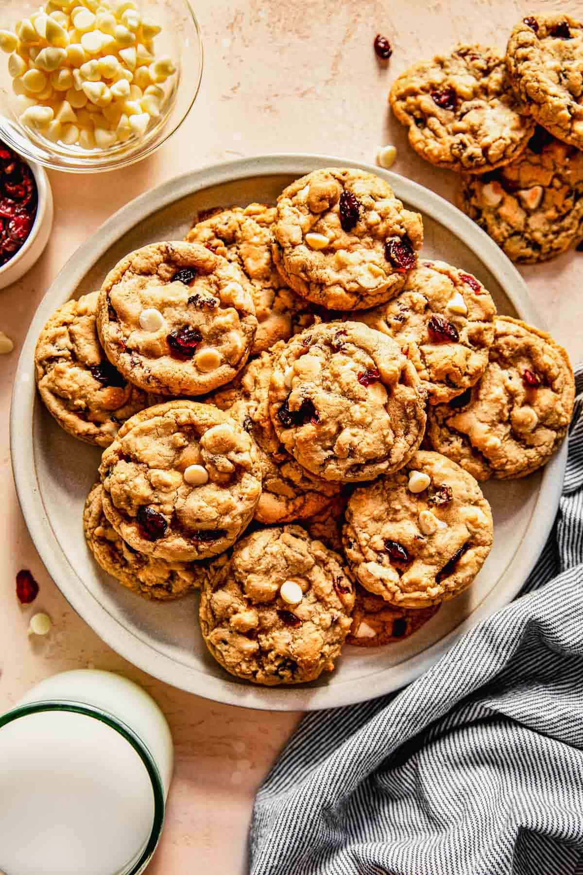 white chocolate and cranberry cookies on a large plate next to a glass of milk and a striped linen towel