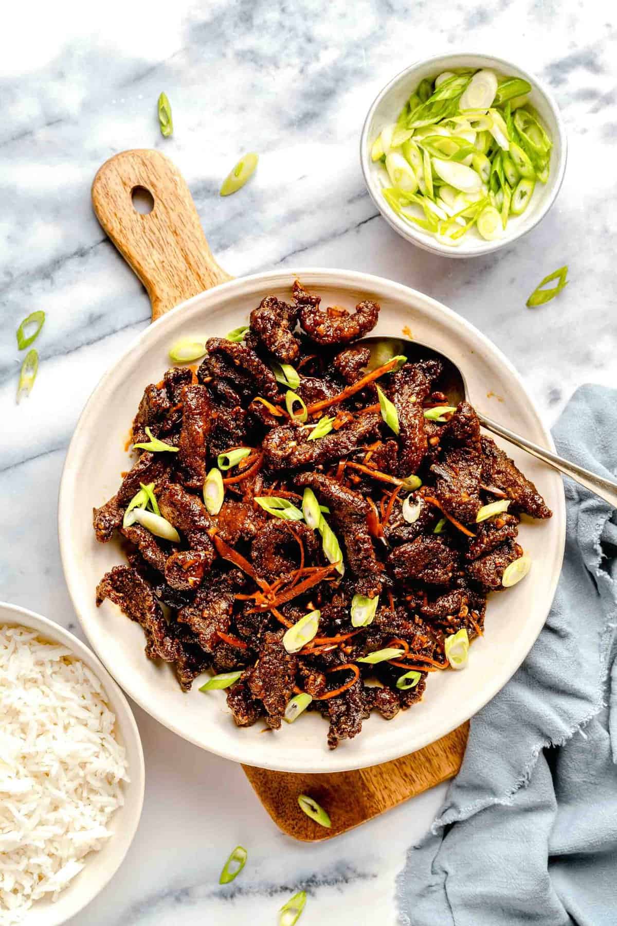 Overhead image of crispy beef on a serving platter near a bowl of rice and a bowl of chopped scallions.