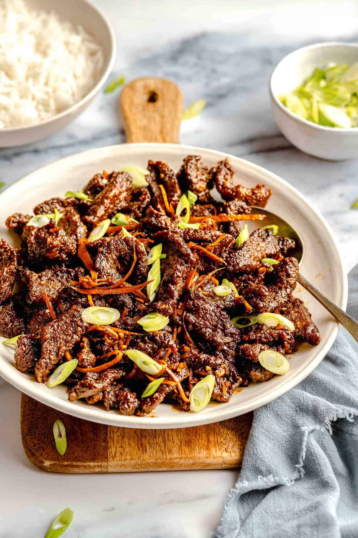 A serving platter of crispy beef on a wooden board near a bowl of rice.