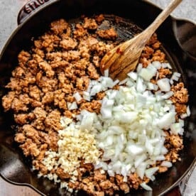 Adding onions and garlic to a skillet full of browned Italian sausage.