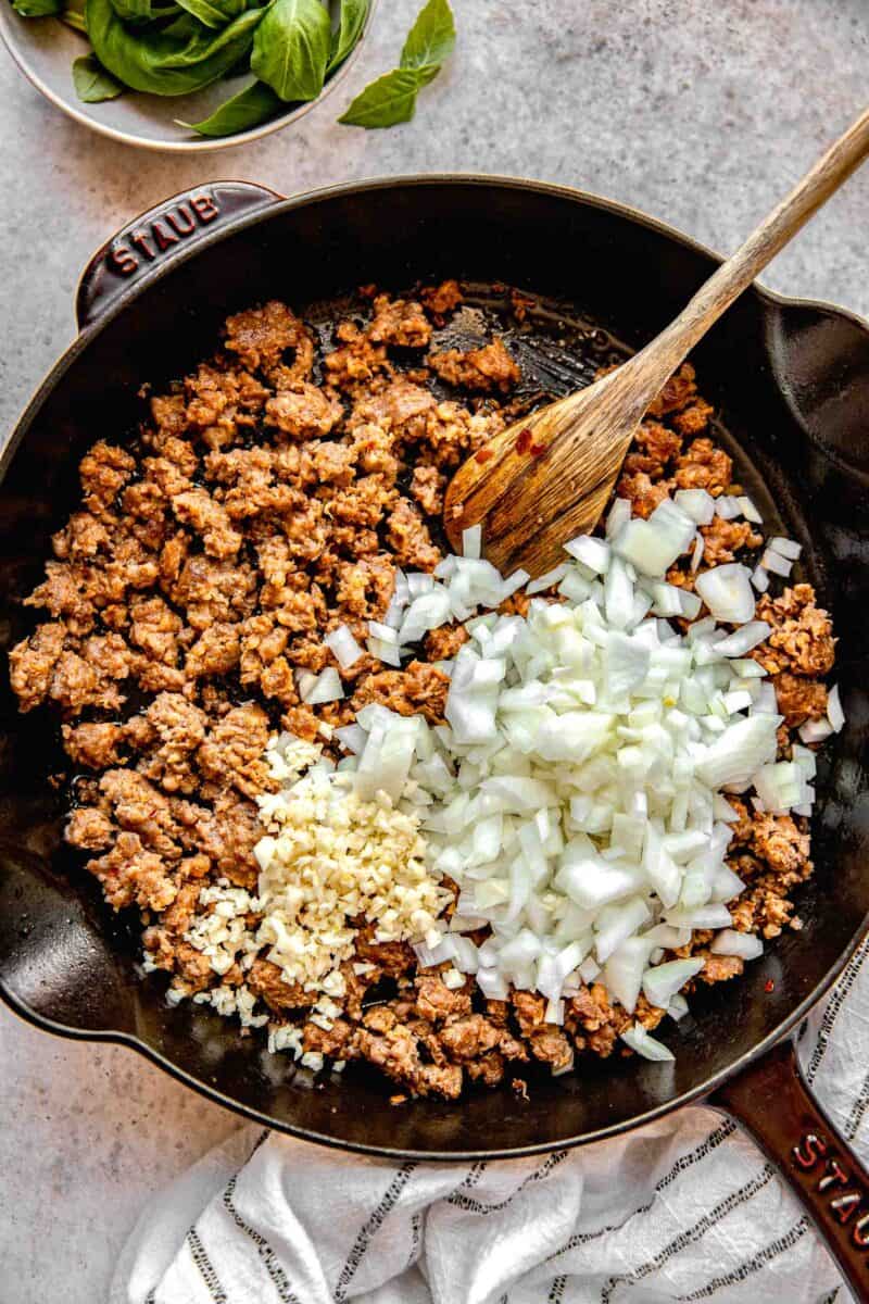 Adding onions and garlic to a skillet full of browned Italian sausage.