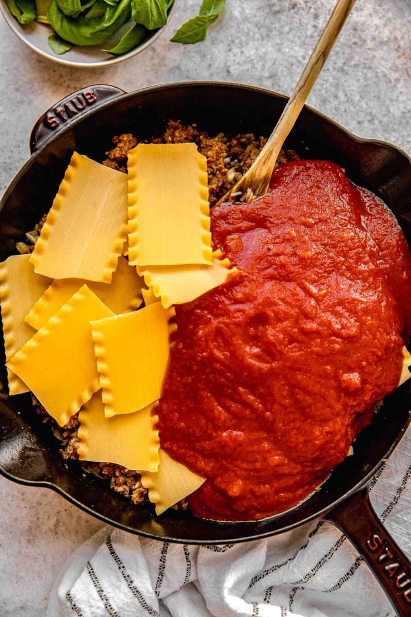 Stirring lasagna noodles and marinara sauce into a skillet with browned sausage, onions, and garlic.