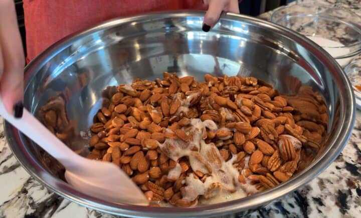 covering raw almonds and pecans in foamy egg white and maple syrup mixture in a large metal bowl