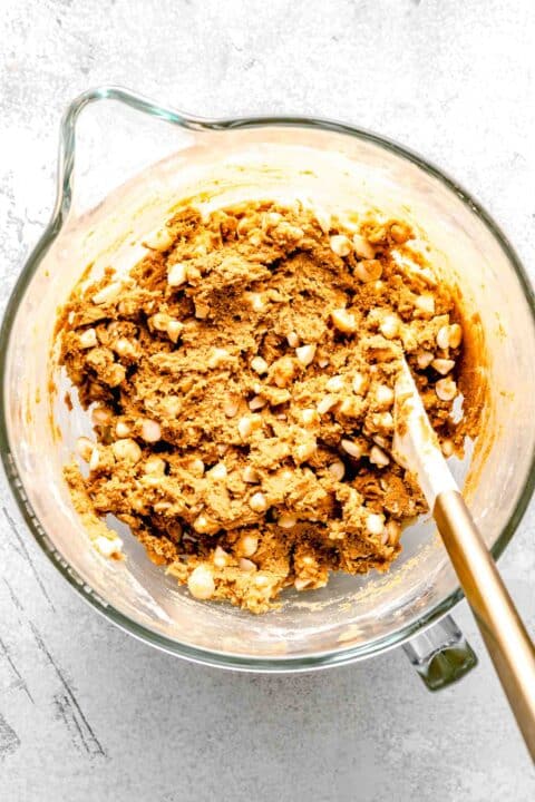 white chocolate and macadamia nuts mixed into the cookie batter in a clear stand mixer bowl with white spatula