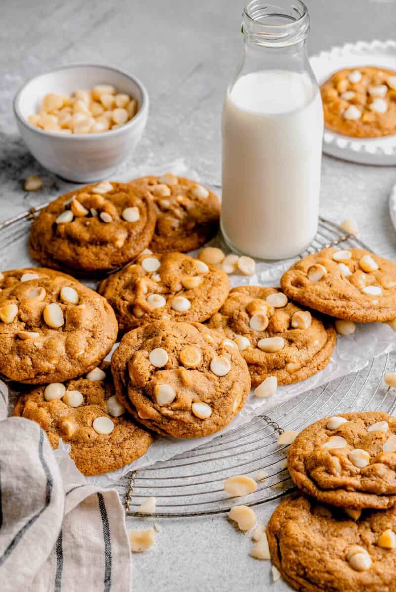 white chocolate macadamia nut cookies on a wire circular rack next to a bottle of milk and bowl of macadamia nuts and striped linen towel