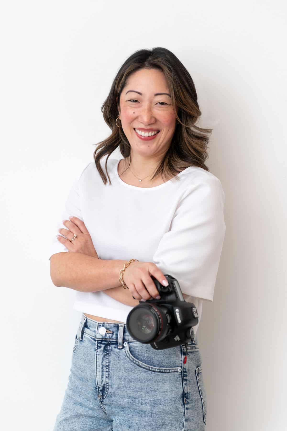 Julie Chiou in a white shirt and light blue jeans holding a camera with arms crossed and smiling against a wall