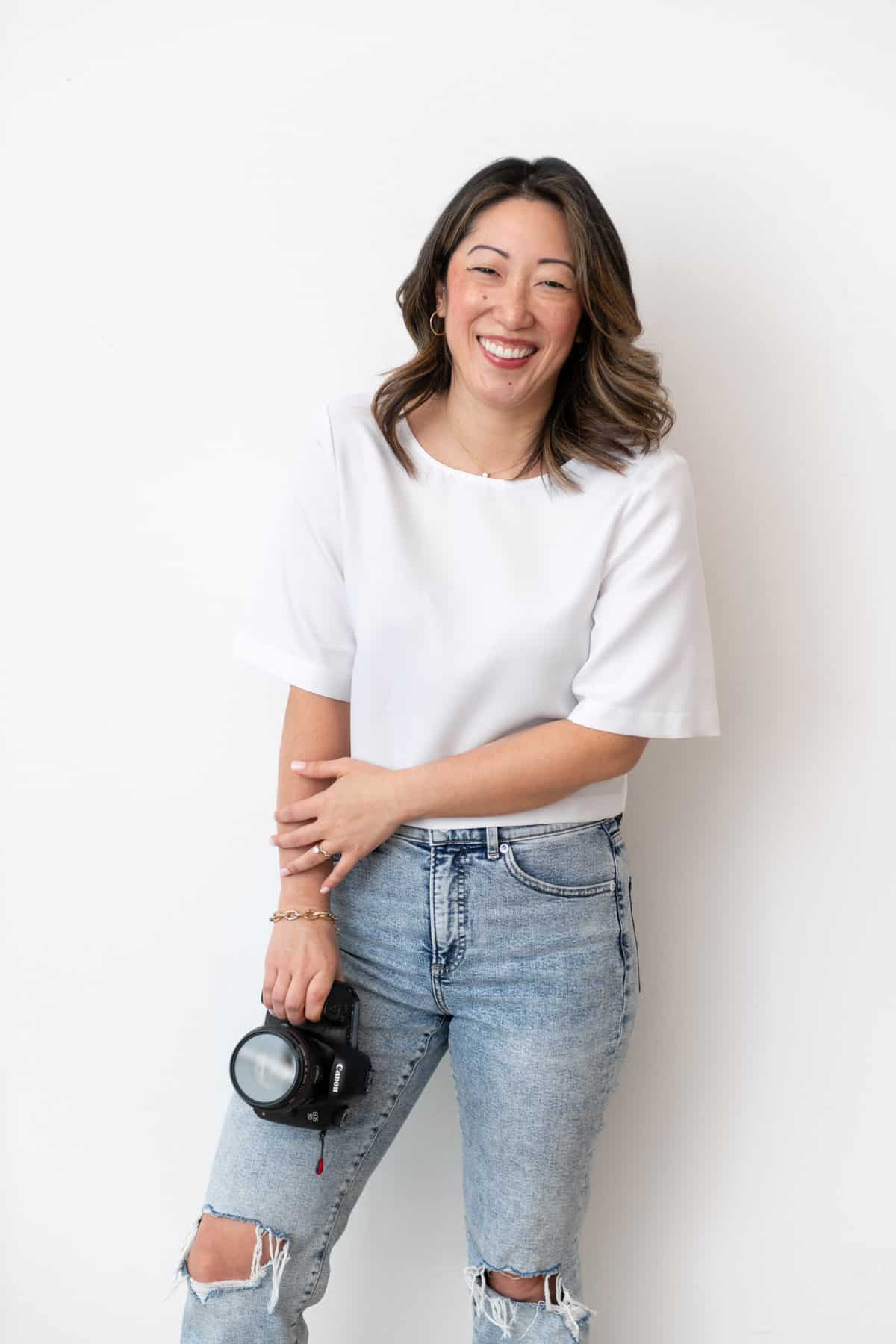 Julie Chiou in a white shirt and light blue jeans holding a camera touching one arm and smiling against a wall