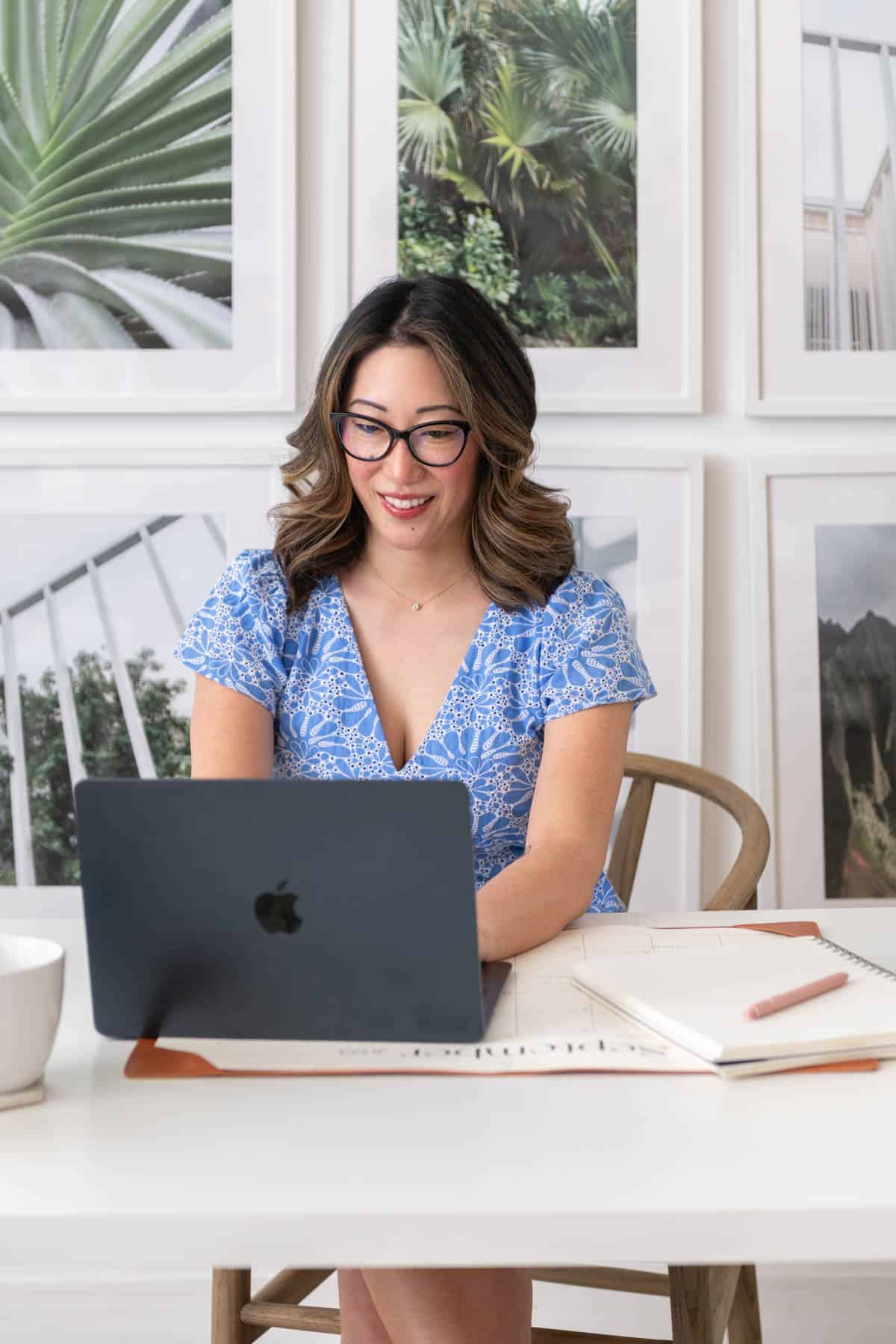 Julie Chiou in a light blue eyelet dress at a desk typing on a black laptop with various framed plant photos in the background