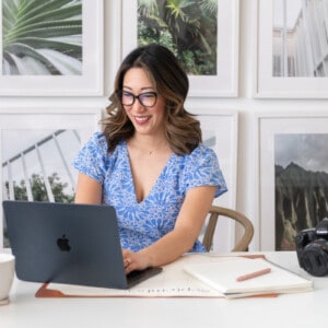 Julie Chiou in a light blue eyelet dress at a desk typing on a black laptop next to her camera and a coffee mug and papers