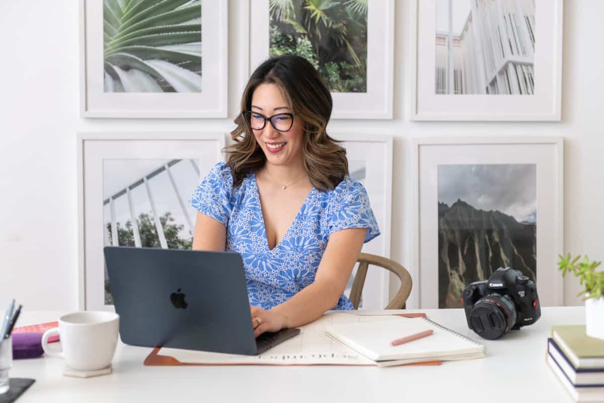 Julie Chiou in a light blue eyelet dress at a desk typing on a black laptop next to her camera and a coffee mug and papers