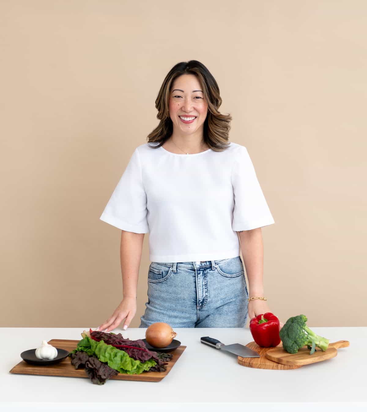 Julie Chiou in a white shirt and light blue jeans standing at a white table with various ingredients and a knife on a cutting board