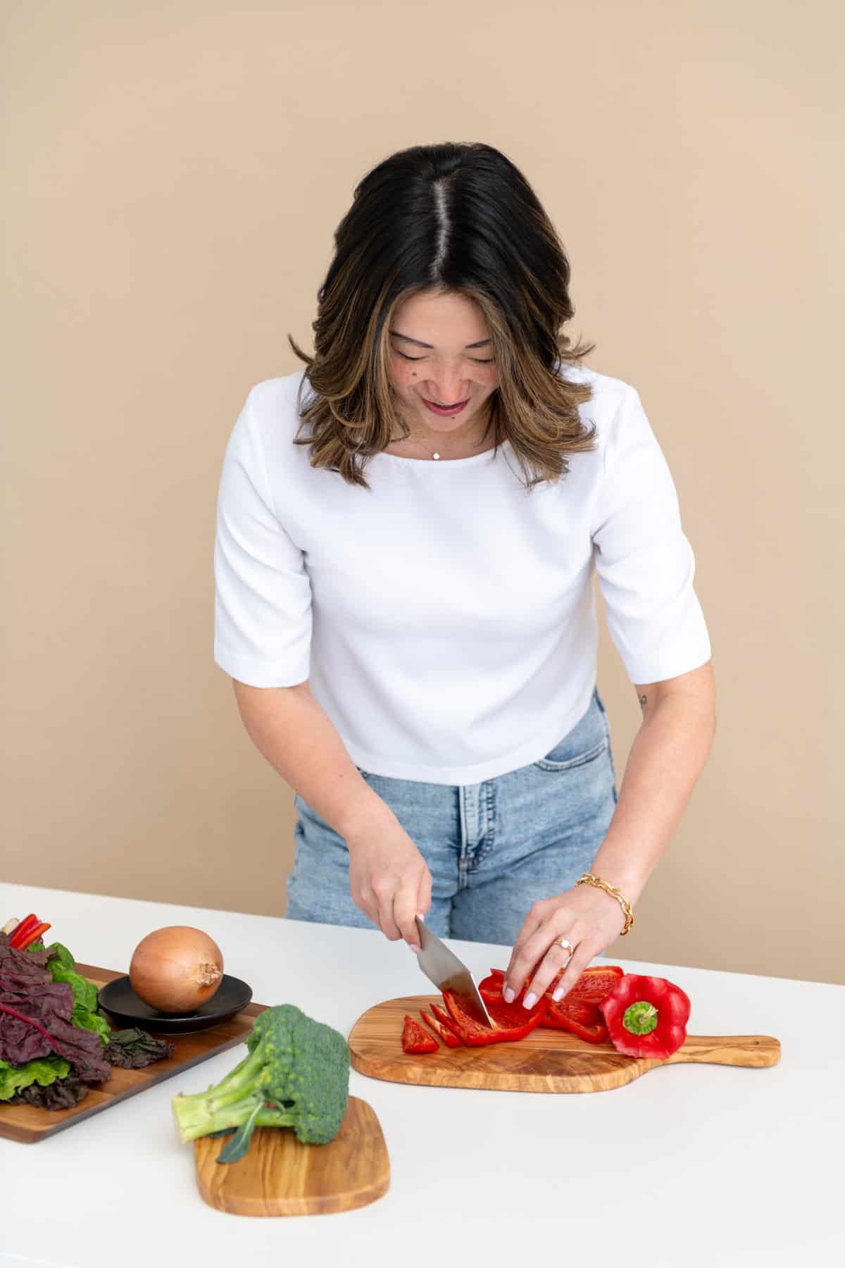 Julie Chiou with white shirt and light blue jeans slicing a red bell pepper on a wooden cutting board