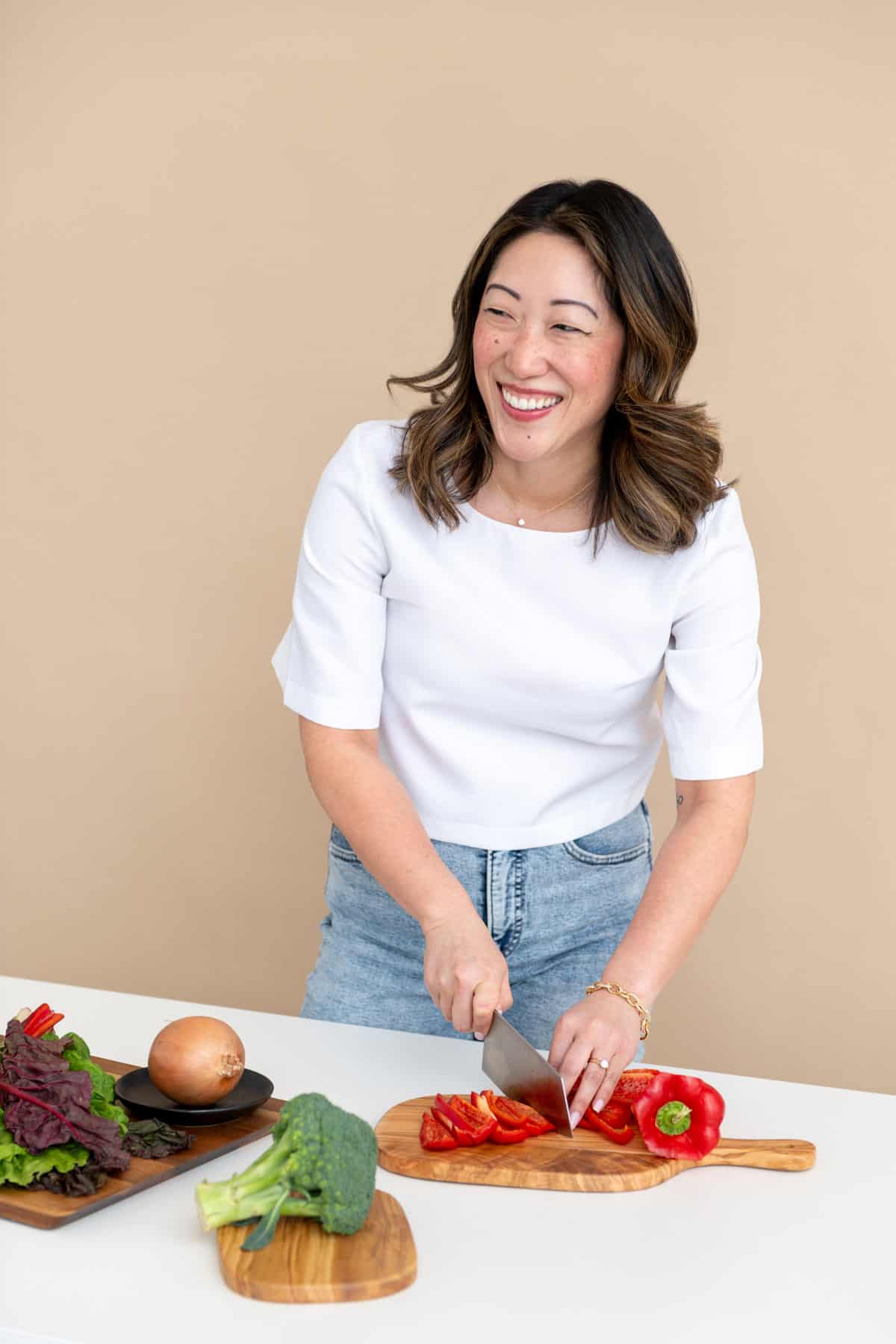 Julie Chiou with white shirt and light blue jeans smiling as she is slicing a red bell pepper