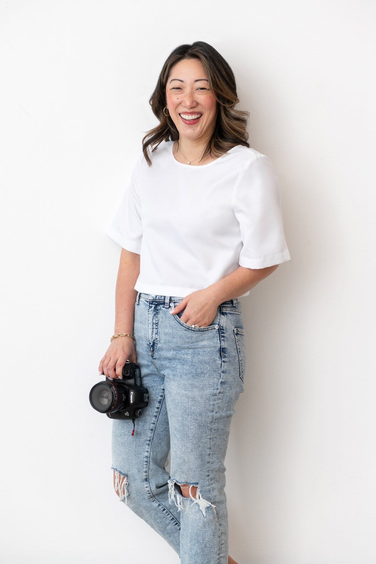 Julie Chiou in a white shirt and light blue jeans holding a camera and smiling against a wall
