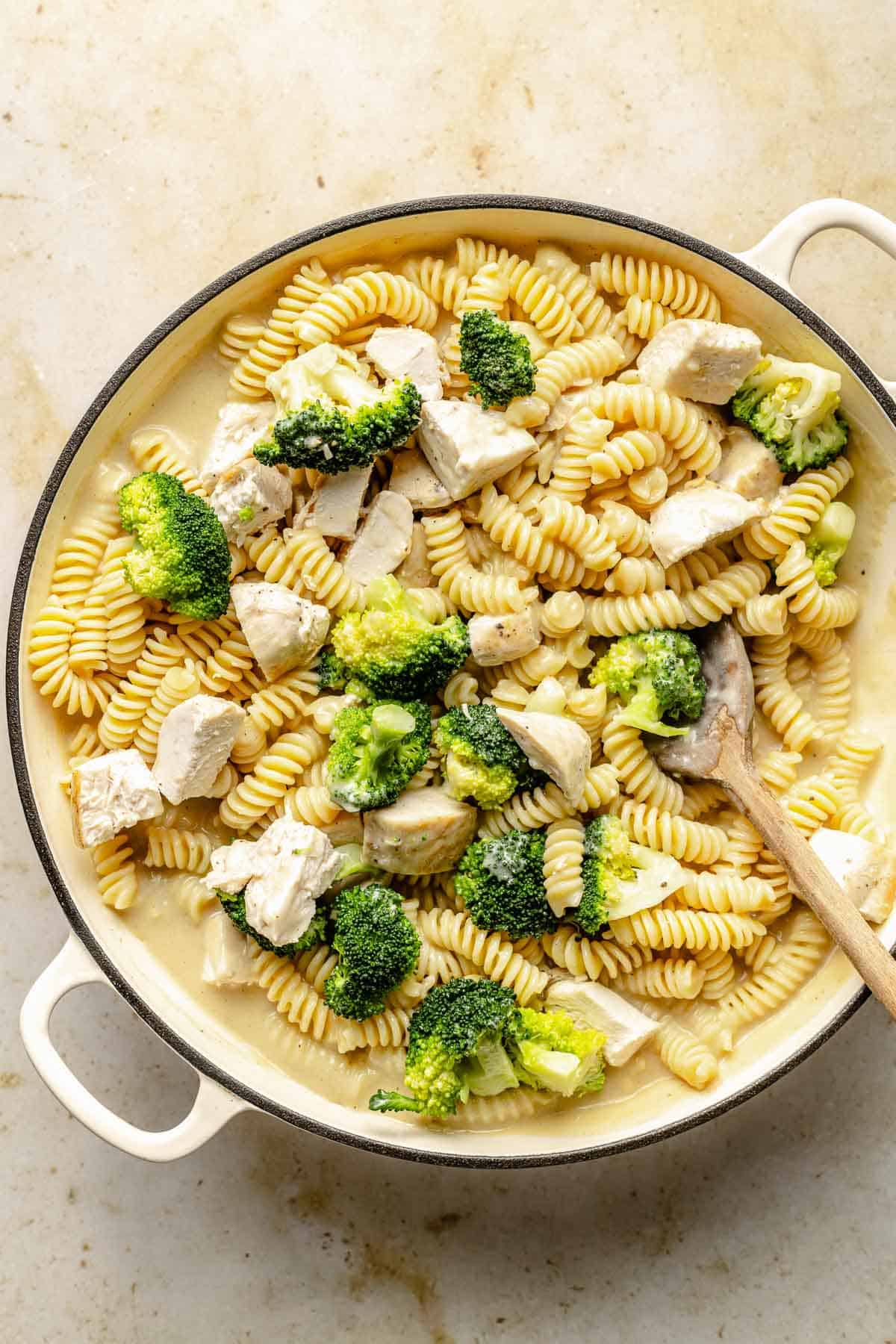 Adding chicken, broccoli, and pasta to Alfredo sauce in a pan.