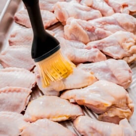 raw chicken wings on a metal baking sheet being brushed with oil with a silicone brush