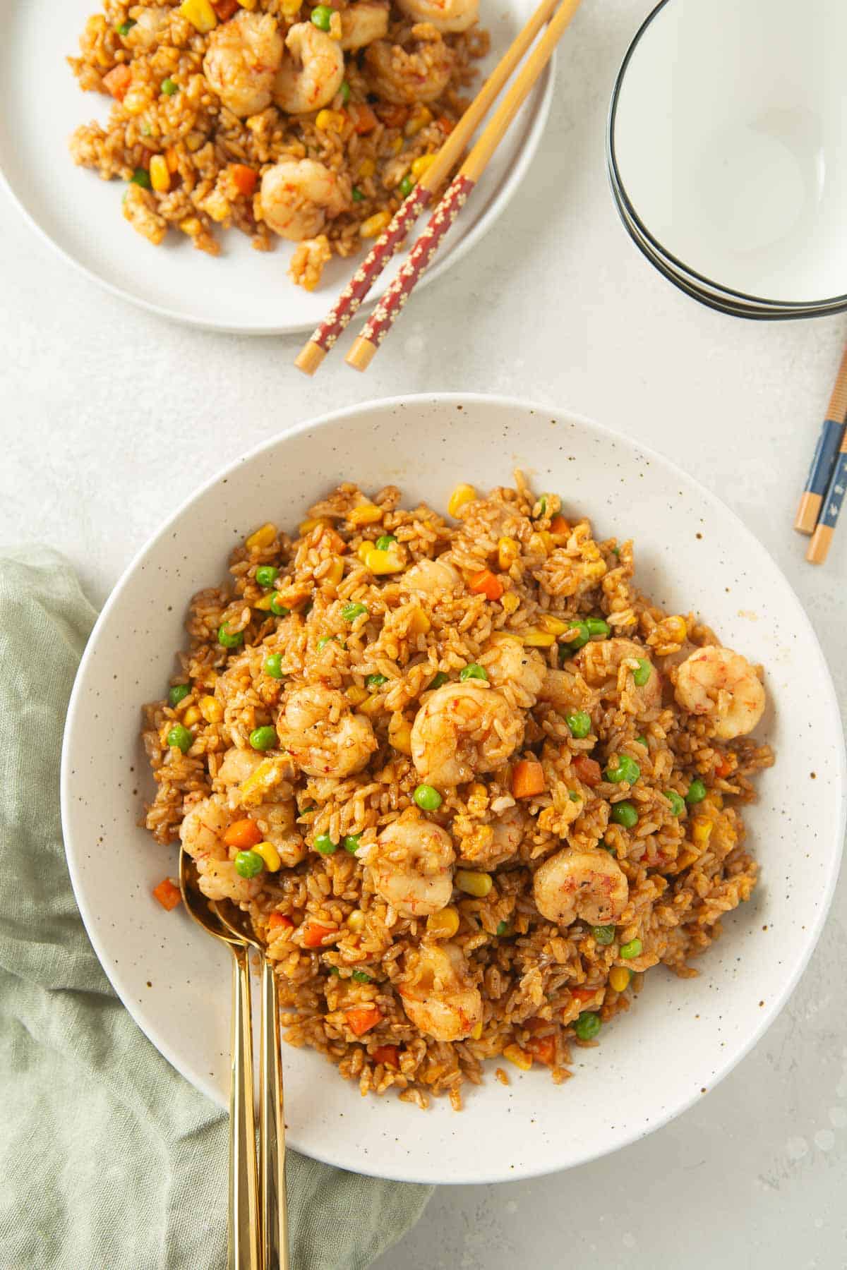 overhead of one large bowl of shrimp fried rice next to a plate of fried rice and wooden chopsticks, empty bowls, and green linen towel