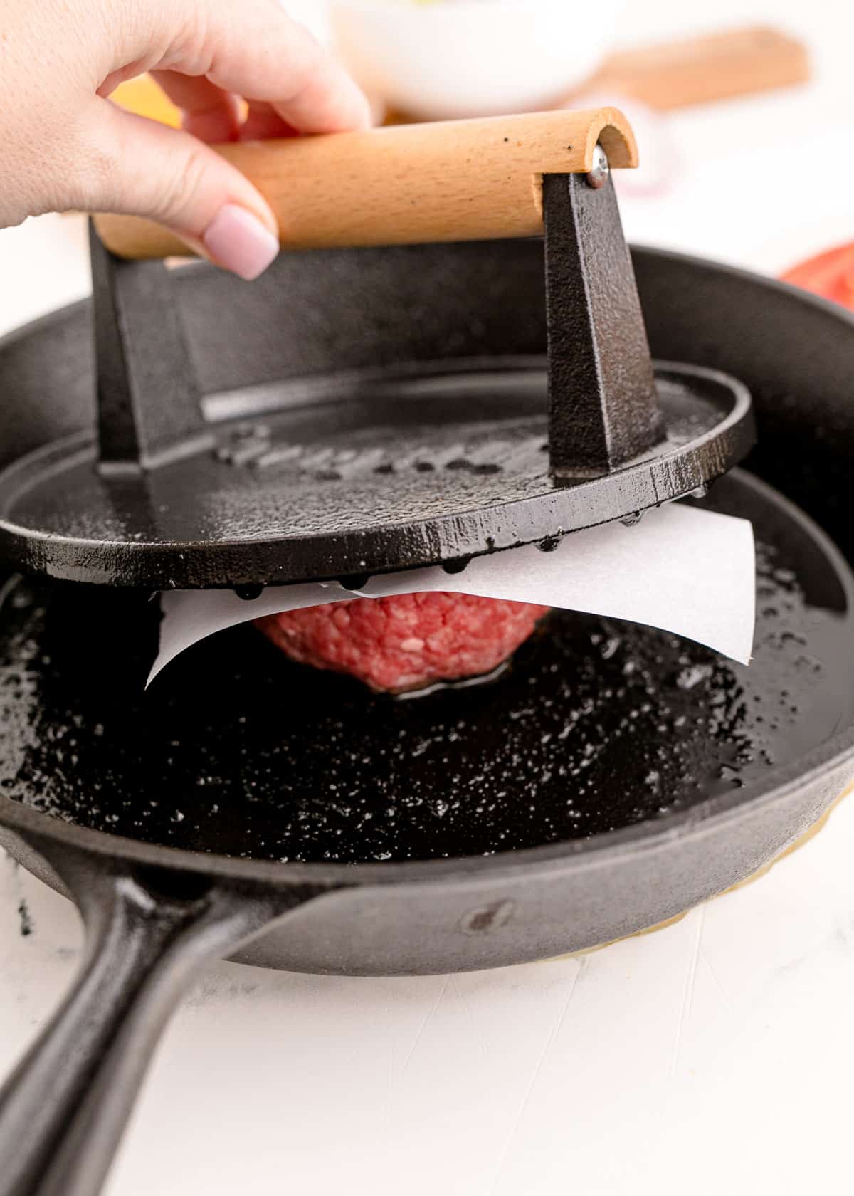 a heavy cast iron meat mallet is being pressed onto a piece of parchment paper on the ball of ground beef in the cast iron skillet